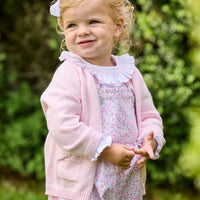 Little English traditional children's clothing. Light pink and green floral overall with ruffle detailing layered over white knit smocked Madeline Blouse in light pink. Light pink cotton bow pocket cardigan added on top.