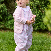 Little English traditional children's clothing. Light pink and green floral overall with ruffle detailing layered over white knit smocked Madeline Blouse in light pink. Light pink cotton bow pocket cardigan added on top.