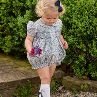 Little English classic children's clothing.  Dressy peter pan bubble for baby girls in blue Wisteria Floral with puff sleeves, ruffle details, and smocking at chest. Paired with white cotton knee high socks with navy bow embroidery