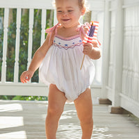 Little English baby girl's smocked bubble for Summer. Smocking with blue stars at center of chest and red seersucker gingham ties at shoulder for 4th of July.