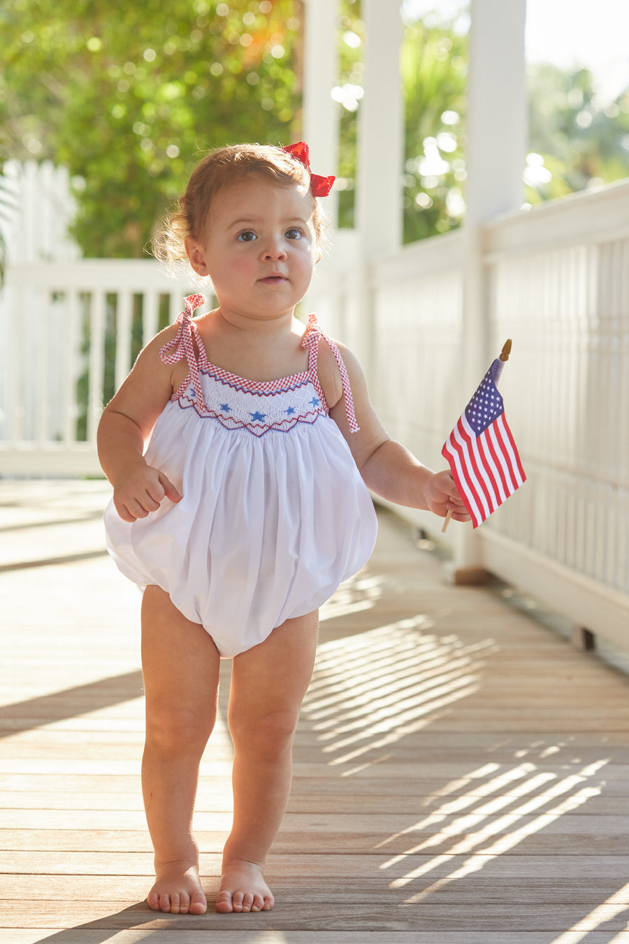 Little English baby girl's smocked bubble for Summer. Smocking with blue stars at center of chest and red seersucker gingham ties at shoulder for 4th of July.